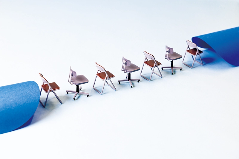 青い紙がめくれたような隙間に、ミニチュアの事務椅子と折りたたみ椅子が一列に並んでいる様子の写真